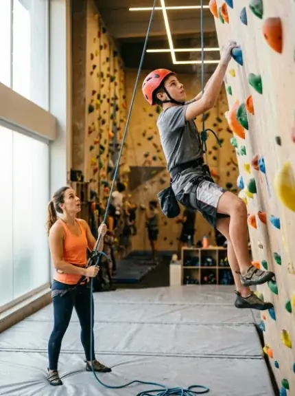 Instrutora fazendo a segurança na corda enquanto um aluno de capacete sobe a parede de escalada